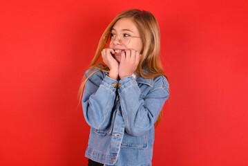 Terrified blonde little kid girl wearing denim jacket over red background looks empty space home alone moonless night