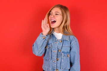 blonde little kid girl wearing denim jacket over red background look empty space holding hand near...