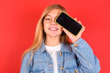 Fototapeta premium blonde little kid girl wearing denim jacket over red background holding modern smartphone covering one eye while smiling