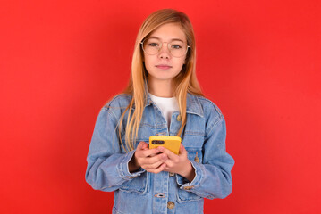 Portrait of serious confident blonde little kid girl wearing denim jacket over red background holding phone in two hands