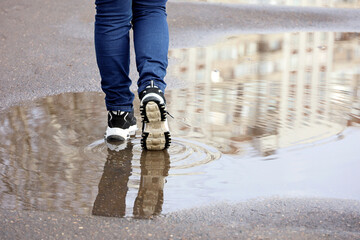 Female legs in jeans and sneakers with reflection in puddle on a street. Rain in city, melting snow...