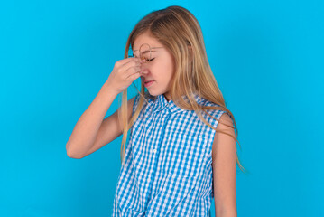 Very upset, little kid girl with glasses wearing plaid shirt over blue background  touching nose between closed eyes, wants to cry, having stressful relationship or having troubles with work