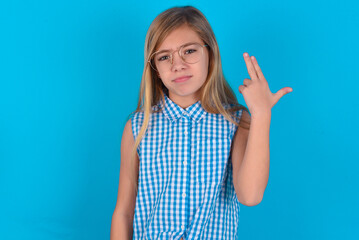 Unhappy little kid girl with glasses wearing plaid shirt over blue background  imitates gun shoot...