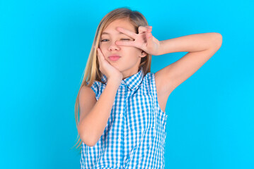 little kid girl with glasses wearing plaid shirt over blue background   making v-sign near eyes. Leisure lifestyle people person celebrate flirt coquettish concept.