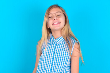 little kid girl with glasses wearing plaid shirt over blue background  with broad smile, shows white teeth, feeling confident rejoices having day off.