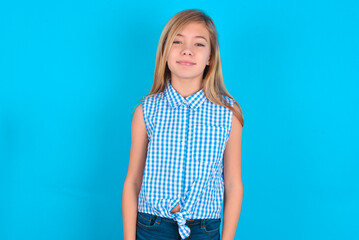 Happy little kid girl with glasses wearing plaid shirt over blue background  looking at camera with charming cute smile.