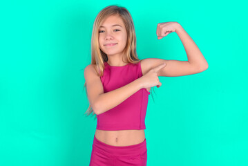 Smiling blonde little kid girl wearing pink sport clothes over green background raises hand to show...