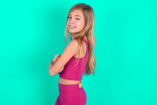 Image of cheerful blonde little kid girl wearing pink sport clothes over green background with arms crossed. Looking and smiling at the camera. Confidence concept.