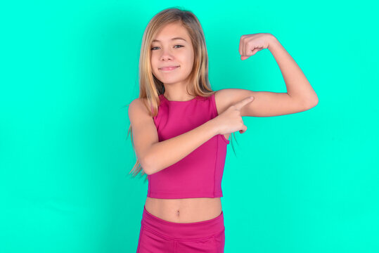 Smiling blonde little kid girl wearing pink sport clothes over green background raises hand to show muscles, feels confident in victory, strong and independent.