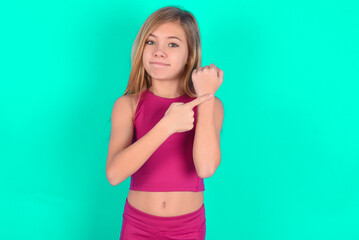 Fototapeta na wymiar blonde little kid girl wearing pink sport clothes over green background in hurry pointing to watch time, impatience, upset and angry for deadline delay