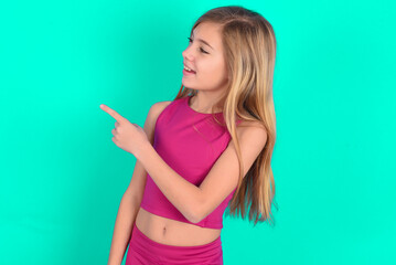 Happy cheerful smiling blonde little kid girl wearing pink sport clothes over green background looking and pointing aside with hand. Copy space and advertisement concept.