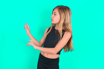 Displeased blonde little kid girl wearing black sport clothes over green background keeps hands towards empty space and asks not come closer sees something unpleasant