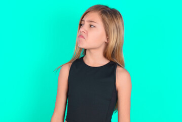 Dissatisfied blonde little kid girl wearing black sport clothes over green background purses lips and has unhappy expression looks away stands offended. Depressed frustrated model.