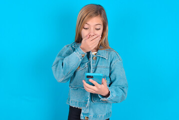 blonde little kid girl wearing denim jacket over blue background being deeply surprised, stares at...