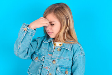Sad blonde little kid girl wearing denim jacket over blue background suffering from headache...