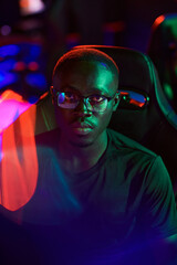 Portrait of serious young black esports participant in eyeglasses sitting in computer chair in neon lights