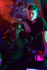 Young man leaning on computer chair with his African-American friend in modern computer club in neon light