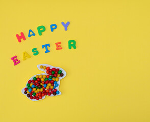 Bright small chocolate candy on Easter bunny shaped plate and phrase Happy Easter made of bright letters on yellow background. Happy Easter concept. Flat lay style with copy space