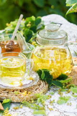 Linden flowers tea in a glass cup and teapot on a white napkin outdoors, yellow drink of linden flower with linden blossoms honey in nature background, healthcare and healthy eating concept