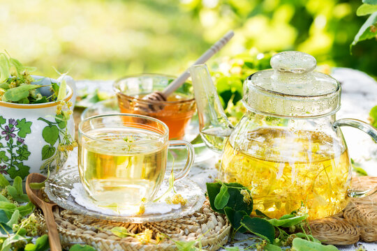 Linden flowers tea in a glass cup and teapot on a white napkin outdoors, yellow drink of linden flower with linden blossoms honey in nature background, healthcare and healthy eating concept