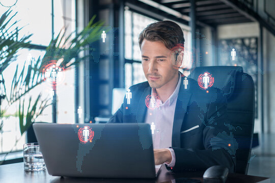 Handsome businessman in suit at workplace working with laptop to hire new employees for international business consulting. HR, social media hologram icons over office background