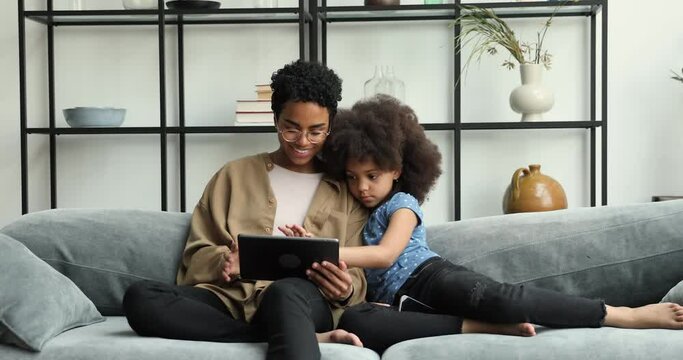 Interested Black preteen girl sit on cozy sofa snuggle up to caring mom nanny grown elder sister work on digital touchpad gadget together with adult person. Happy mom child play games online on pad pc