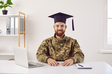 Happy bearded young soldier student in camouflage uniform and graduate hat sitting at table with...