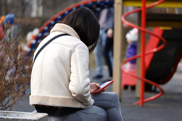Girl in white coat and jeans sitting with a smartphone on the bench on playground in spring park....