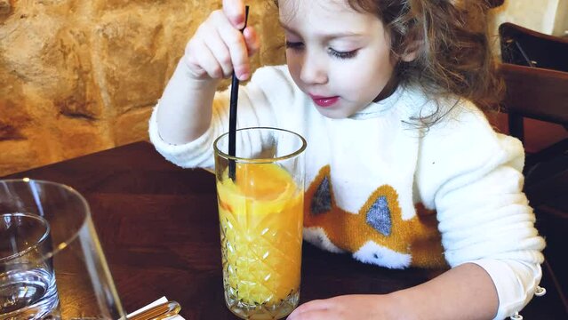 Cute young girl drinking freshly squeezed orange juice from a glass with a straw in a restaurant
