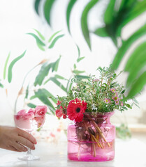 Women hand holding champagne glass with rose petals and flower ice cube on table with flowers bunch in pink glass vase on white table with blurred branch background. Refreshing summer drink.Front view
