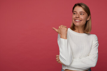 indoor portrait of young female posing over red background points with a finger aside at copy space and laugh