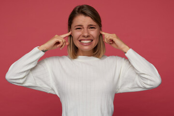 Fototapeta premium indoor portrait of young female posing over red background closed her ears with fingers, irritated and angry