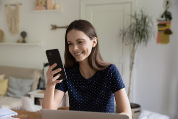 Young woman sit at table with laptop holds modern smartphone smile read received message with pleasant news, learn new software use mobile application, working or studying alone at home, tech concept