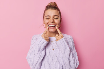 Young glad woman points at toothy smile shows perfect white teeth being in good mood keeps eyes closed dressed in casual knitted jumper isolated over pink background. Positive emotions concept