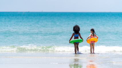 Little African and Asian girl in swimwear and swim ring playing in sea water together at tropical...