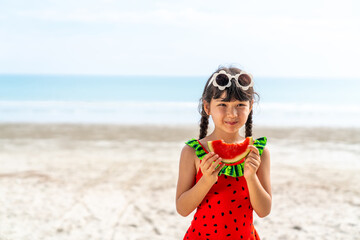 Little Asian child girl in swimwear eating watermelon while playing with family on tropical beach...