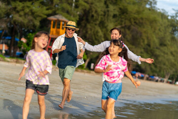 Happy Asian family father and mother with two little daughter walking and playing together on the beach at summer sunset. Parents with child girl kid enjoy and fun outdoor lifestyle on summer vacation
