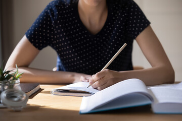 Unknown student girl sit at table with textbook, holds pencil writes essay, prepare for university exams, studying alone indoor, language practice, cram, learns subject, makes chapter overview concept