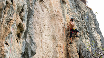 Confidence young Asian man climber with safety rope climbing on rocky mountain at tropical island in sunny day. Strong handsome male enjoy outdoor active lifestyle and extreme sport on summer vacation