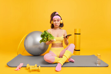 Displeased Asian woman dressed in sportswear leads healthy lifestyle holds lettuce salad and...