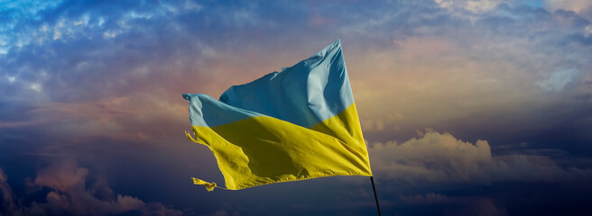 Flag of Ukraine on dramatic cloudy sky for wide banner or billboard about the war. Damaged Ukrainian flag. The national symbol of Ukraine in the struggle for independence, freedom and sovereignty.