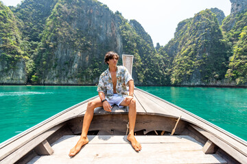 Fototapeta na wymiar Portrait of Young handsome Asian man sitting on the boat passing beach lagoon in summer sunny day. Healthy guy relax and enjoy outdoor lifestyle on travel vacation at tropical island in Thailand