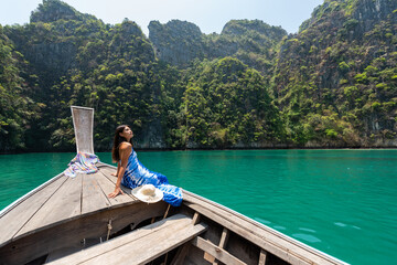 Young beautiful Asian woman in blue dress sitting on the boat passing island beach lagoon in summer sunny day. Happy female relax and enjoy outdoor lifestyle together on summer vacation in Thailand