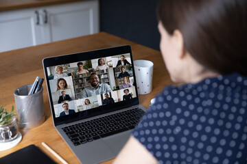 Faces of diverse young business people take part in video conference, laptop screen over female shoulder sit at table involved in group virtual meeting. Modern tech, video call, communication concept