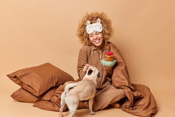 Glad young woman plays with pug dog poses crossed legs wears nightwear eats delicious pancakes for breakfast enjoys company of favorite pet sits under blanket isolated over beige background.