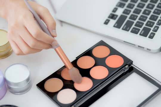 Hands of woman with learning makeup with brush on cheek on laptop computer with tutorial course online, female teaching and explain make up with makeup palette and cosmetic on streaming social media.