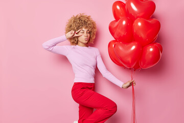 Romantic young woman keeps lips folded makes peace gesture over eye has fun prepares for holiday holds bunch of heart helium balloons wears casual jumper and trousers isolated over pink wall