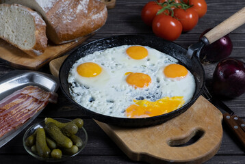 rustic fried eggs on a dark wooden table with tomatoes, a glass of vodka, beacon, bread and pickled gherkins. in a dark key