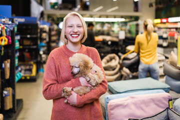 Beautiful young blond woman buying accessories and food for her poodle puppy in pet shop.