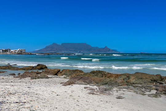 Beach, rocks and ocean with Table Mountain in the background. Against a clear blue sky. 
